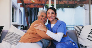 An elderly woman and a home care nurse in blue scrubs embracing and smiling while sitting on a couch in a cozy outdoor setting. This image represents compassionate caregiving and the impact of home care marketing in building strong client relationships.