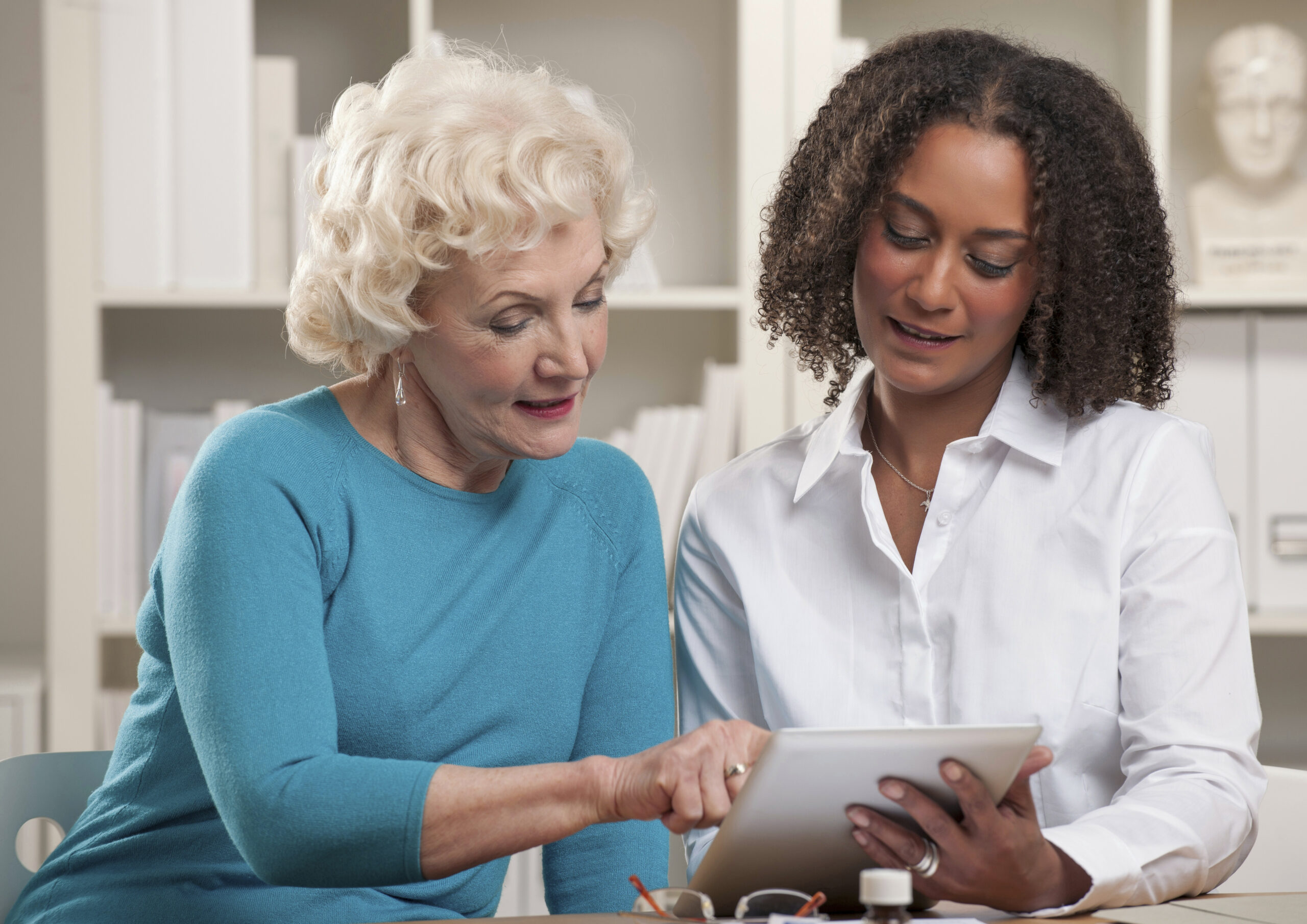 An elderly woman in a teal sweater and a younger caregiver in a white shirt discussing home care services while using a digital tablet. A professional setting with bookshelves in the background, representing effective home care marketing strategies through digital engagement.