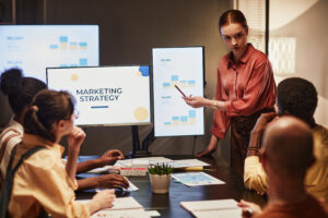 A young businesswoman in an orange shirt presents a marketing strategy to her team in an office. Screens display charts and analytical data supporting her presentation.