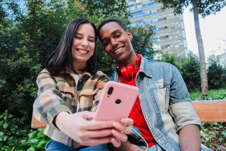 Young couple sitting on a bench, smiling while watching short-form videos on a smartphone.
