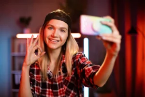 Young woman recording a selfie video indoors, showing an OK hand gesture under colorful lights.