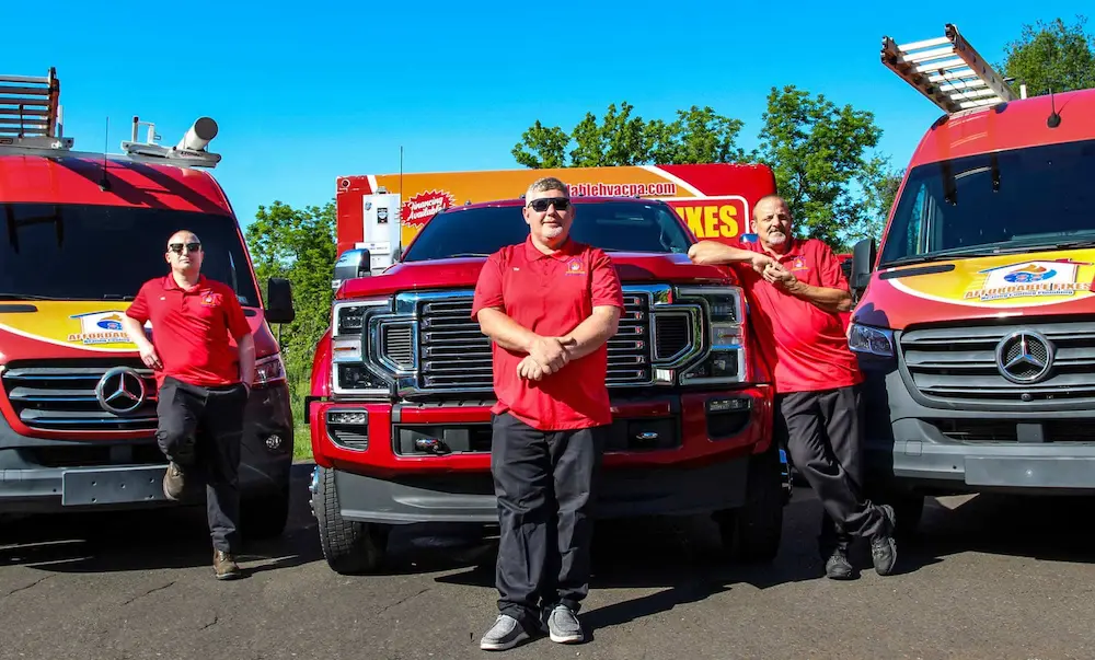 Two men in red shirts stand cheerfully beside a large red truck with a vibrant advertising banner, set against a backdrop of green trees.