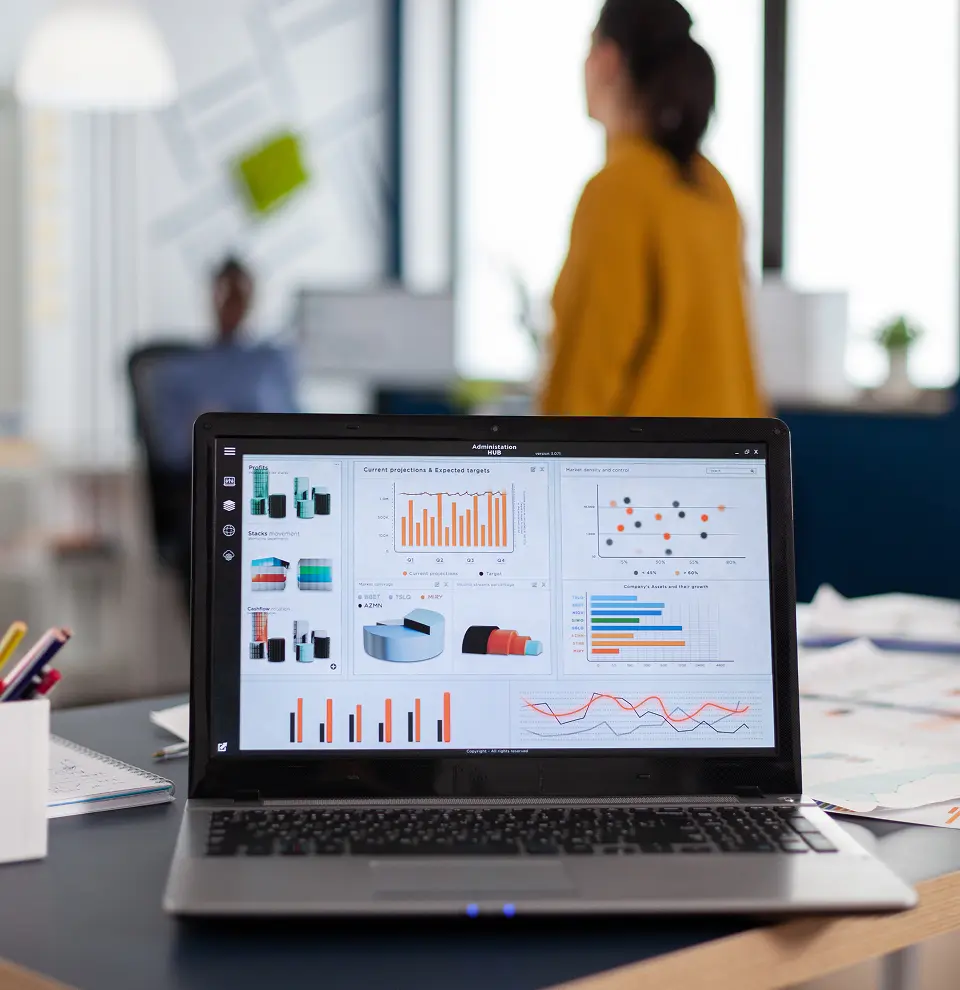 Laptop displaying business analytics dashboards and charts, with an office environment in the background.