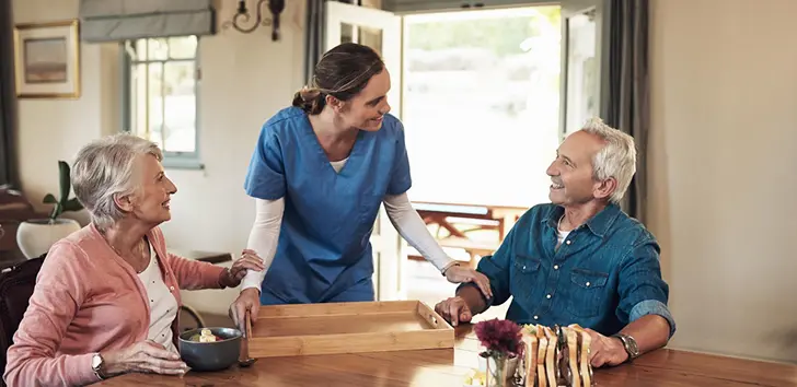 Caregiver in blue scrubs serving a meal tray to two smiling elderly people at a dining table