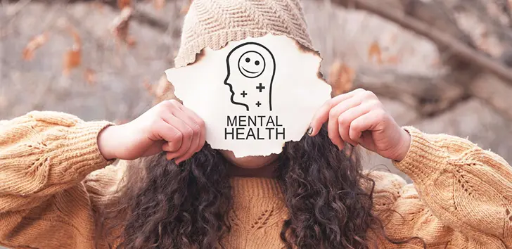 Woman holding a torn paper sign with a mental health icon and the words "Mental Health" in front of her face outdoors