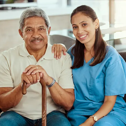 Smiling caregiver in blue scrubs sitting beside an elderly man holding a cane