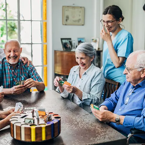 Caregiver in blue scrubs watching over a group of cheerful elderly people playing cards at a table