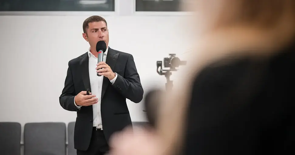 A man in a black suit speaking into a microphone at a business presentation or conference event
