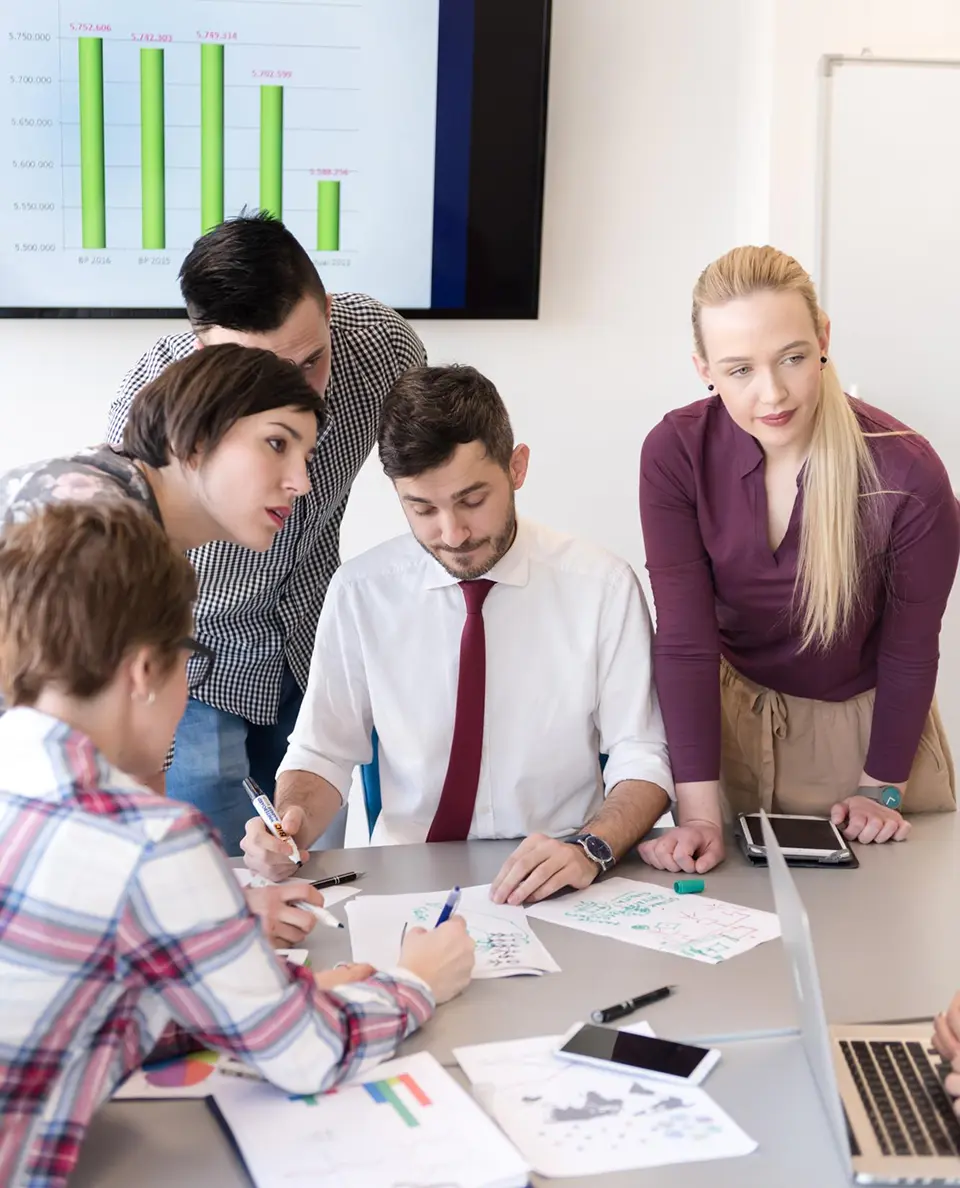 Business team gathered around a conference table reviewing documents with a bar chart on a screen behind them